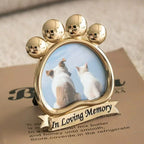 Shiny gold metal pet picture frame in the shape of a paw. Engraved with 'In Loving Memory' on the base. Pictured holding a photo of a dog and cat sitting together. A high-quality, freestanding keepsake for pet owners to display on a desk or shelf.