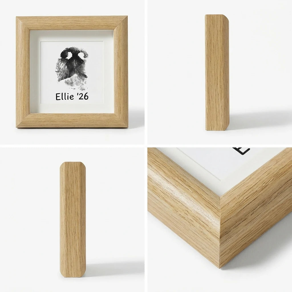 Bright, minimalist lifestyle shot of natural wood pet memorial shadow box frame leaning on a light wood surface. Features a clear black dog paw print impression with custom text 'Ellie '26'. Styled with a dried botanical stem for a peaceful, Scandi home decor tribute