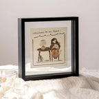 Lifestyle view of a modern black floating frame standing on a textured table. Displays a charming pet illustration reading 'Welcome to our home'. The square, double-glass design creates a see-through border, perfect for displaying art or photos