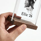 Close-up detail view of a hand holding the solid dark walnut wood base of the pet memorial stand. The shot highlights the thick natural wood grain texture and the precision slot that securely holds the clear acrylic panel. Demonstrates the premium craftsmanship and sturdy foundation of this tribute.