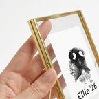 Close-up detail view of a hand holding the corner of the gold brass floating frame. The shot emphasizes the thick, high-quality metal border and the double-pane glass mechanism that securely encases the custom dog nose print on white paper. Showcases the premium construction of this keepsake