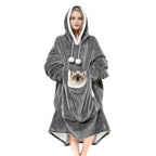 A smiling woman standing in a cozy gray oversized wearable blanket hoodie with a large front kangaroo pouch holding a fluffy Siamese cat. Hands-free pet carrier sweatshirt.