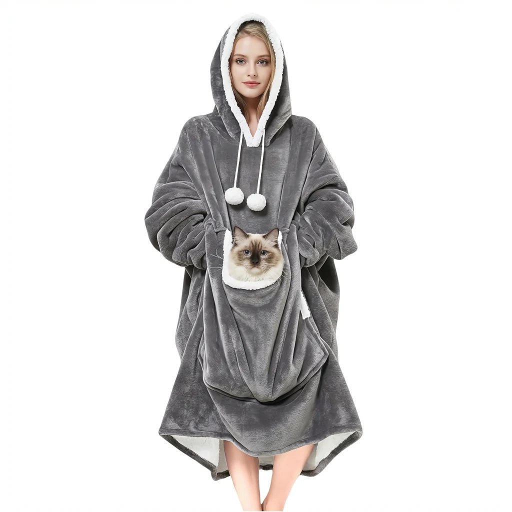 A smiling woman standing in a cozy gray oversized wearable blanket hoodie with a large front kangaroo pouch holding a fluffy Siamese cat. Hands-free pet carrier sweatshirt.