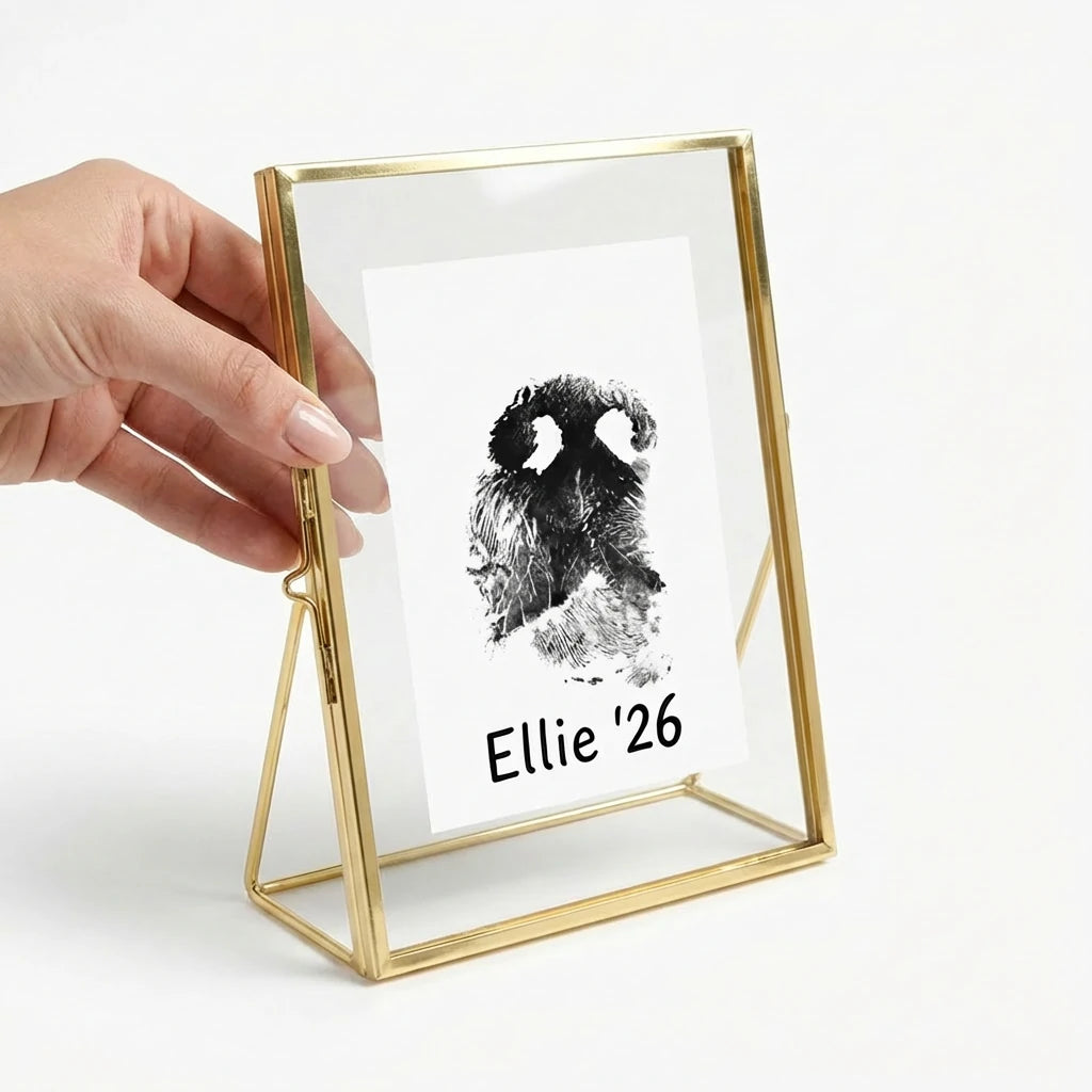 A female hand gently holding the gold brass floating glass pet memorial frame, providing a clear size reference and sense of scale. The frame contains a custom dog nose print. This image conveys the tangible comfort of giving a personalized sympathy gift that a grieving pet owner can hold close to cherish their dog's memory.