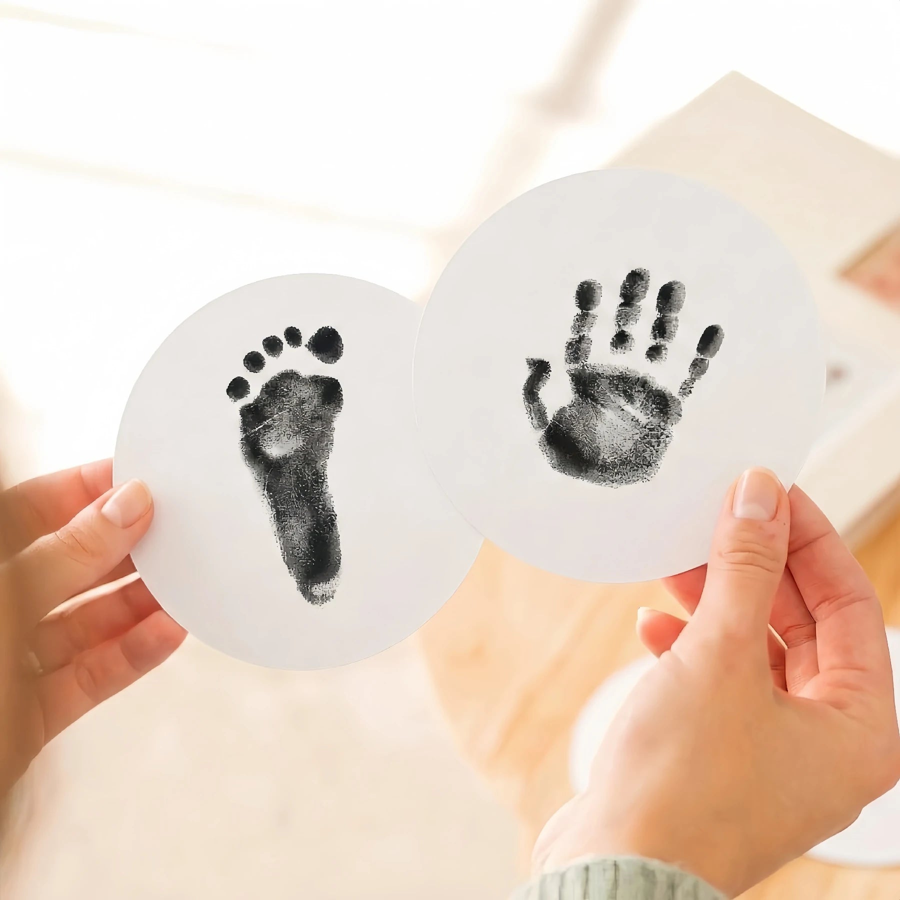 Close-up of a person's hands holding two round paw print keepsake cards featuring a high-contrast black baby footprint and handprint. Demonstrates the detailed results of a black ink print kit for DIY pet memorial goods.