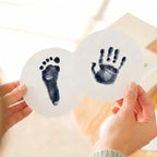 Close-up of a person's hands holding two round paw print keepsake cards featuring a high-contrast navy baby footprint and handprint. Demonstrates the detailed results of a navy ink print kit for DIY pet memorial goods.