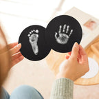 Close-up of a person's hands holding two round paw print keepsake cards featuring a high-contrast white baby footprint and handprint. Demonstrates the detailed results of a silver ink print kit for DIY pet memorial goods.