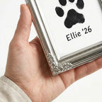 Close-up of a hand holding the corner of the vintage silver pet frame. Highlights the intricate floral filigree detail and the sleek metal rim. A premium way to display a black paw print memorial for a dog or cat.