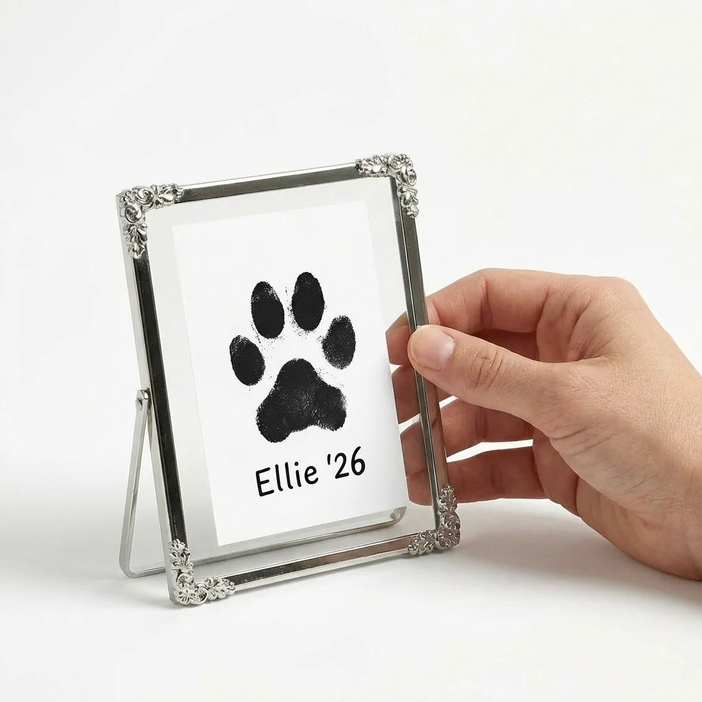 Side angle view of a silver metal frame held by hand. Demonstrates the compact size and the sturdy wire back for easy display. The vintage floral corners frame a beloved pet's paw impression. A sophisticated touch for any home.