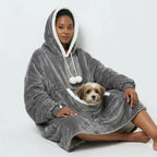 A woman sitting comfortably cross-legged on the floor wearing a grey oversized wearable blanket hoodie. A small fluffy dog is resting inside the large front kangaroo pouch. Cozy fleece loungewear for dog moms.