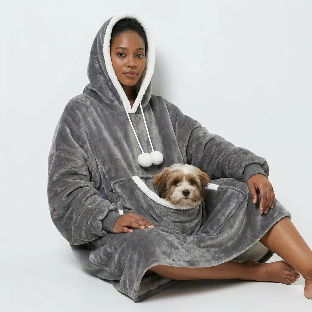 A woman sitting comfortably cross-legged on the floor wearing a grey oversized wearable blanket hoodie. A small fluffy dog is resting inside the large front kangaroo pouch. Cozy fleece loungewear for dog moms.