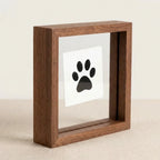 Angled side view of small square walnut wood floating frame featuring a simple black paw print. Highlights deep profile that allows the frame to stand freely on any flat surface. Minimalist double-glass design for modern pet remembrance decor.