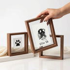 Hand arranging a walnut wood floating frame set to create a pet memorial gallery. Shows placing a rectangular custom nose print frame beside a square paw print frame. Highlights the versatile, see-through design perfect for modern shelf styling.