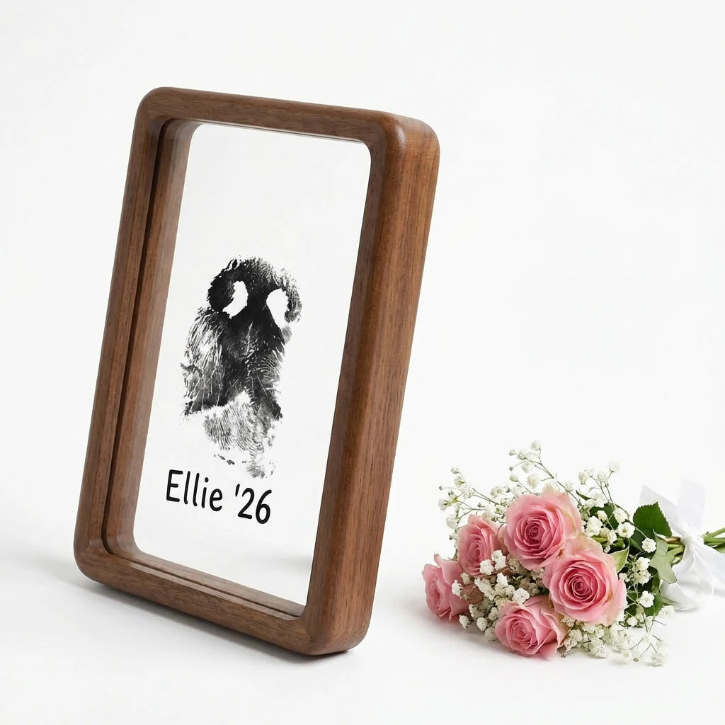 Hand presenting premium walnut wood floating frame with rounded edges. Shows distinct dog nose print (snout print) 'Ellie '26' suspended in double glass. Styled with roses to depict a touching, modern sympathy gift for pet loss remembrance