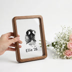 Hand presenting premium walnut wood floating frame with rounded edges. Shows distinct dog nose print (snout print) 'Ellie '26' suspended in double glass. Styled with roses to depict a touching, modern sympathy gift for pet loss remembrance