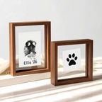 Lifestyle display of matching walnut wood floating frame set. Shows rectangular frame with custom dog nose print (snout print) 'Ellie '26' beside a square frame with classic paw print. Modern double glass design creates a see-through, minimalist pet tribute