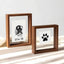 Lifestyle display of matching walnut wood floating frame set. Shows rectangular frame with custom dog nose print (snout print) 'Ellie '26' beside a square frame with classic paw print. Modern double glass design creates a see-through, minimalist pet tribute